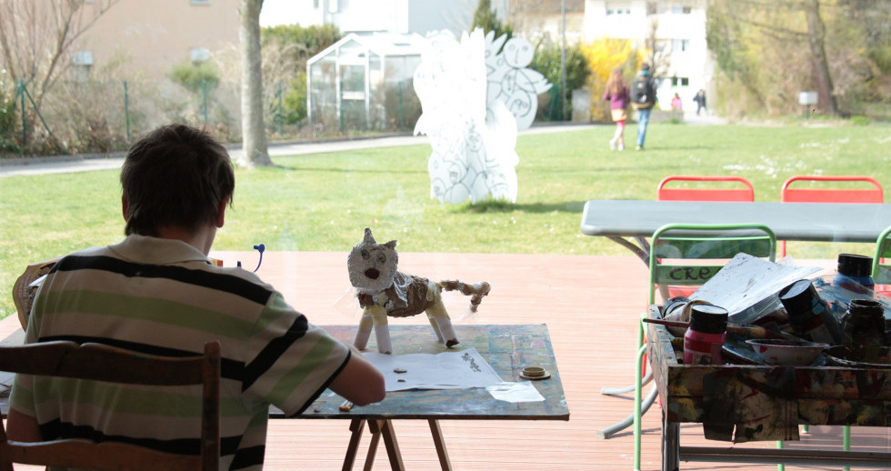 Eine Person aufgenommen von hinten an einem farbverschmierten Arbeitstisch. Darauf eine Katze aus unterschiedlichen Materialien: Klebband, Karton und Papier. Daneben Malwerkzeug. Im Hintergrund der Aussenaufnahme ist eine Wiese, in der Mitte Skulptur.