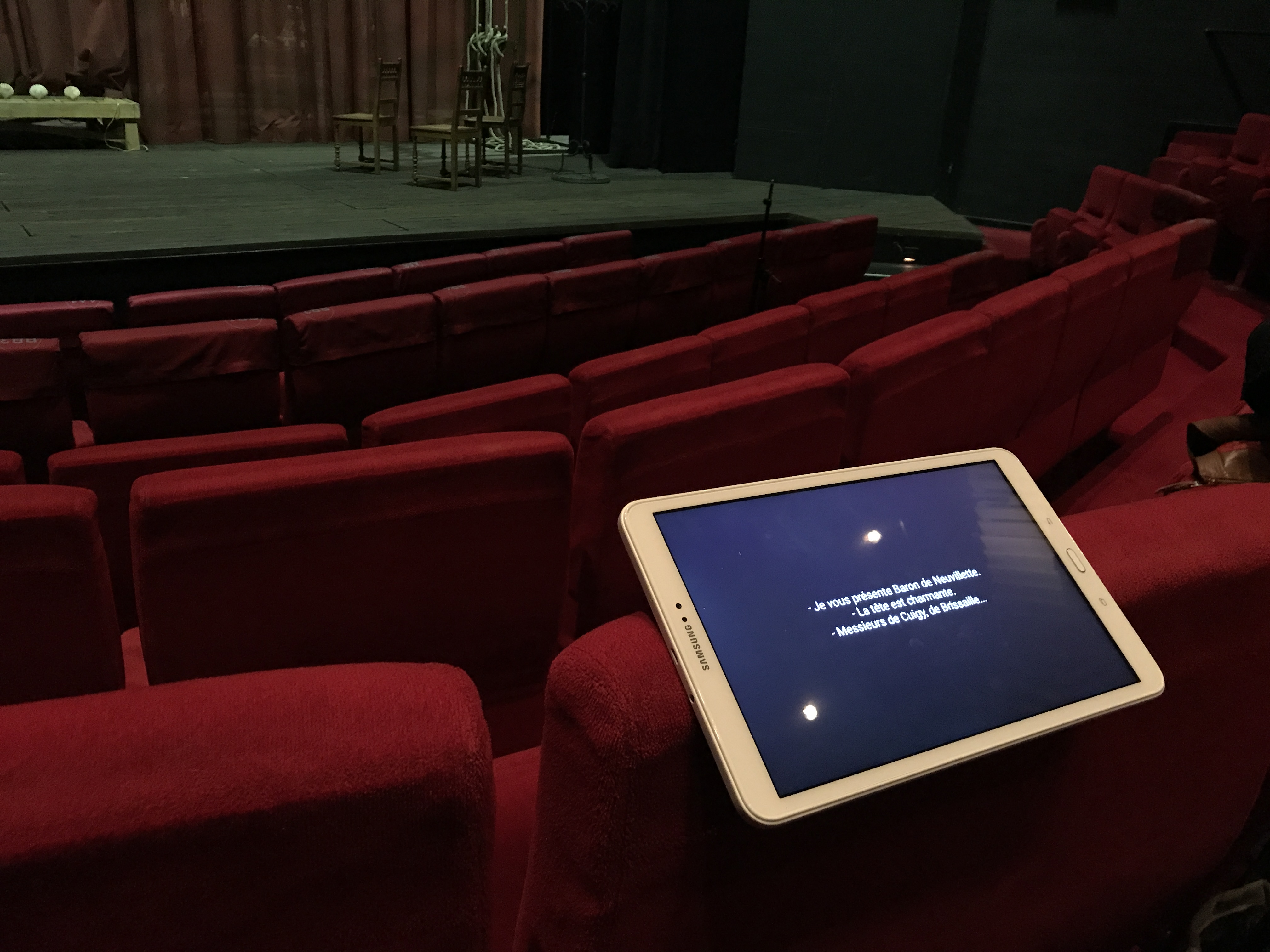 Une tablette posée en équilibre sur le dossier d’un siège en velours rouge dans une salle de théâtre vide. Au se-cond plan, la scène. La tablette est allumée, trois courtes phrases s’affichent sur l’écran.