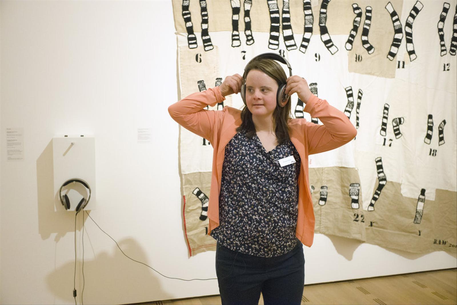 Eine junge Frau steht vor einem grossen Stoffbild. Sie trägt ein Namensschild auf der Bluse und Kopfhörer, die über ein Kabel an einer Vorrichtung an der Wand verbunden sind. Das Bild ist beige-weiss, die «Chromosomen» sind schwarz-weiss gestreift. 