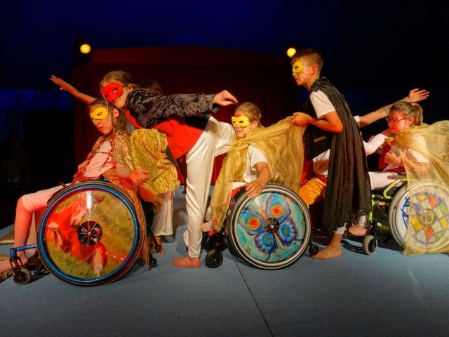 Fünf Kinder in einer Reihe – zwei stehen, drei sitzen im Rollstuhl – bilden einen Zug: Mit ausgestrecktem Arm oder Bein sind sie jeweils zum Kind vor- oder hinter sich verbunden. Sie tragen rote oder gelbe Augenmasken, schwarze oder goldfarbene Umhänge.