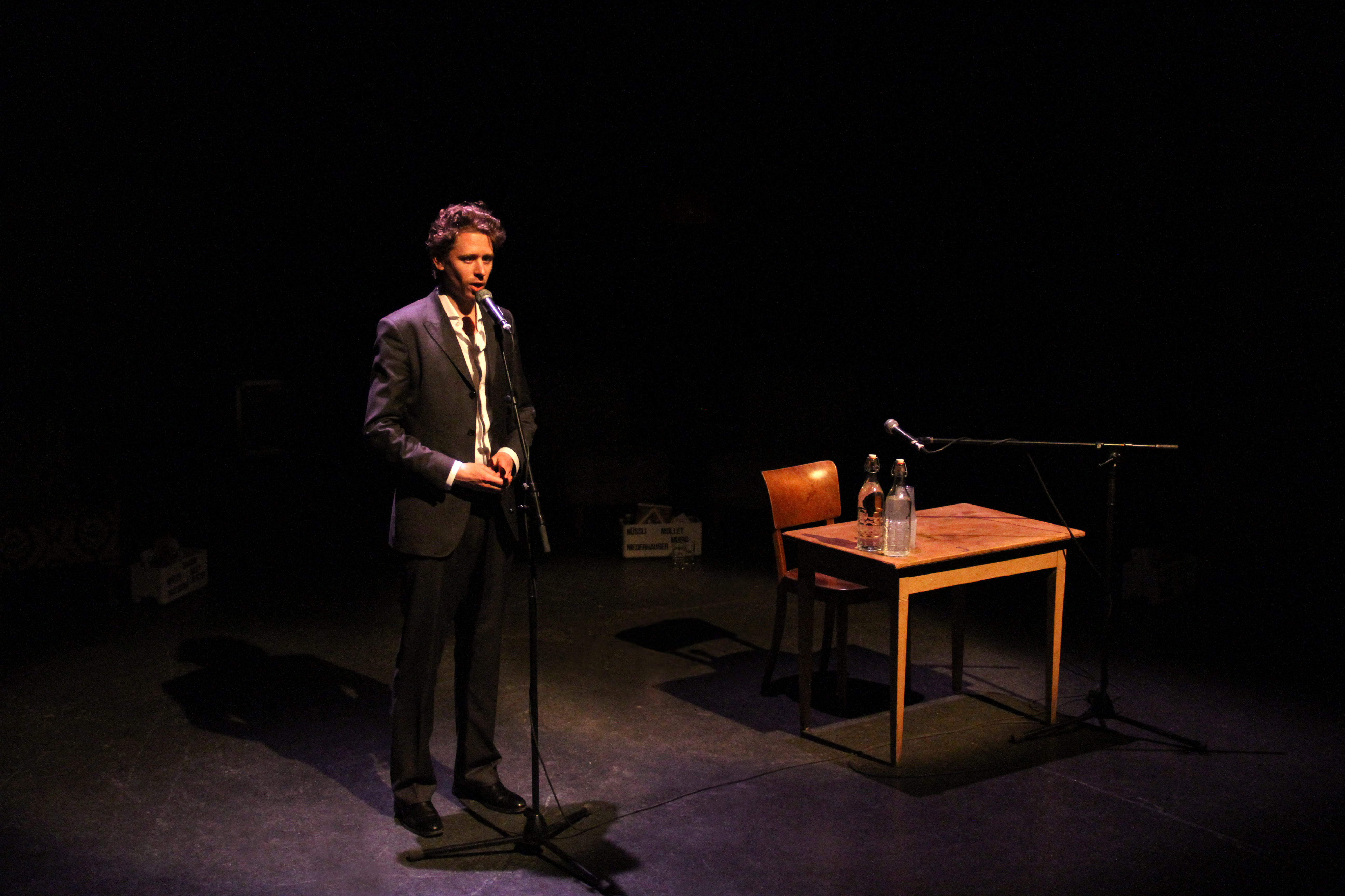 Ein junger Mann in schwarzem Anzug und weissem Hemd steht auf einer Bühne, ein Standmikrofon vor sich. Auf dem Holztischchen rechts von ihm zwei Wasserflaschen, dahinter ein Holzstuhl. Ein Standmikrofon ist auf Sitzhöhe eingestellt. Dunkler Hintergrund.