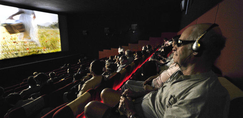 Une salle de cinéma remplie. Au premier plan, une personne de profil avec des lunettes noires et un casque au-dio. Sur l’écran, une personne dans une prairie fleurie, une longue robe blanche et un panier à la main, les sous-titres en bas.