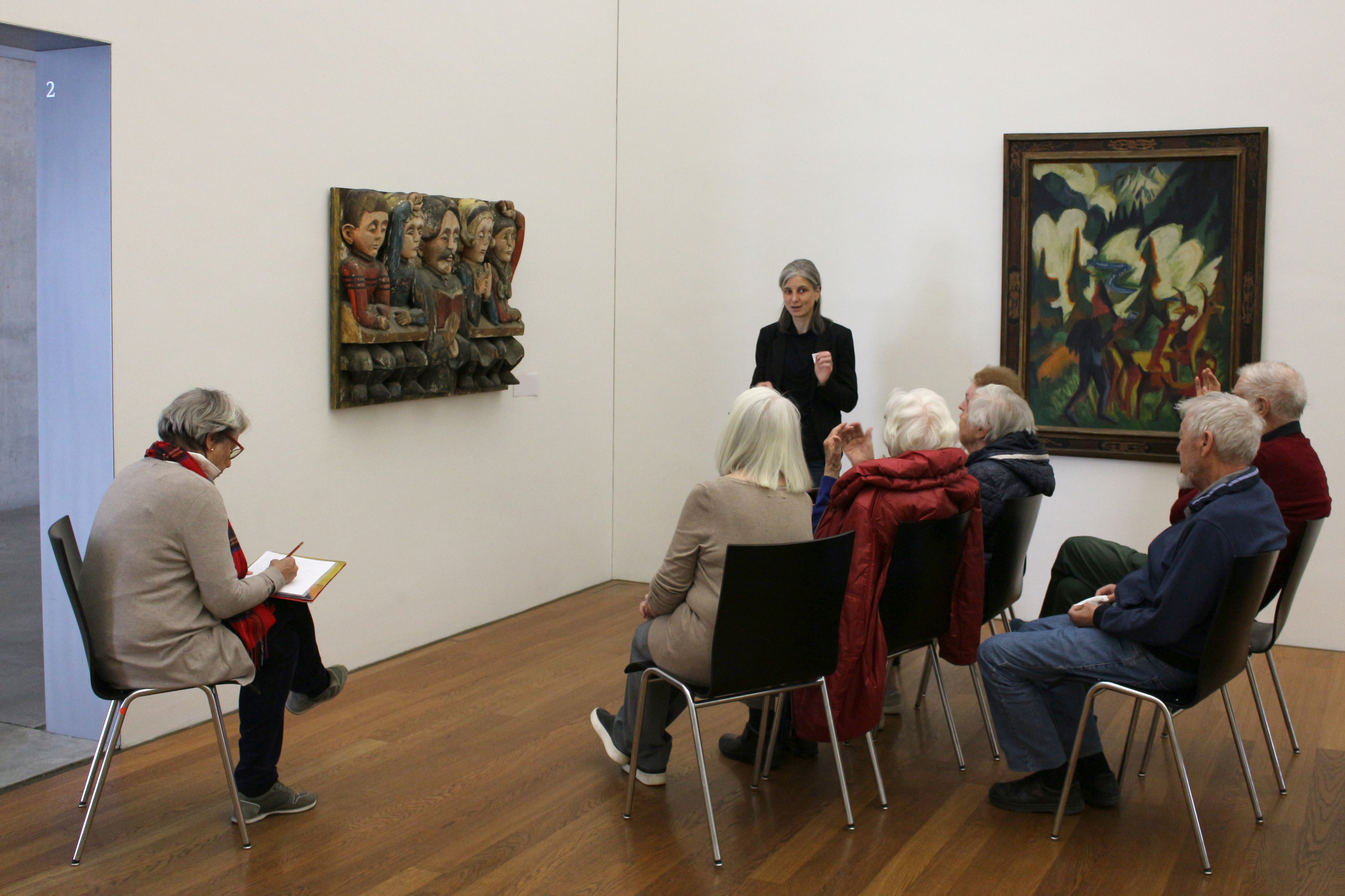 Sechs ältere Menschen sitzen vor einem Holzrelief, aus dem fünf menschliche Figuren den Betrachtenden frontal zugewandt sind. Eine Frau steht neben dem Relief und blickt zu den Sitzenden. Etwas abseits sitzt eine Frau, die auf ein Papierblatt schreibt.