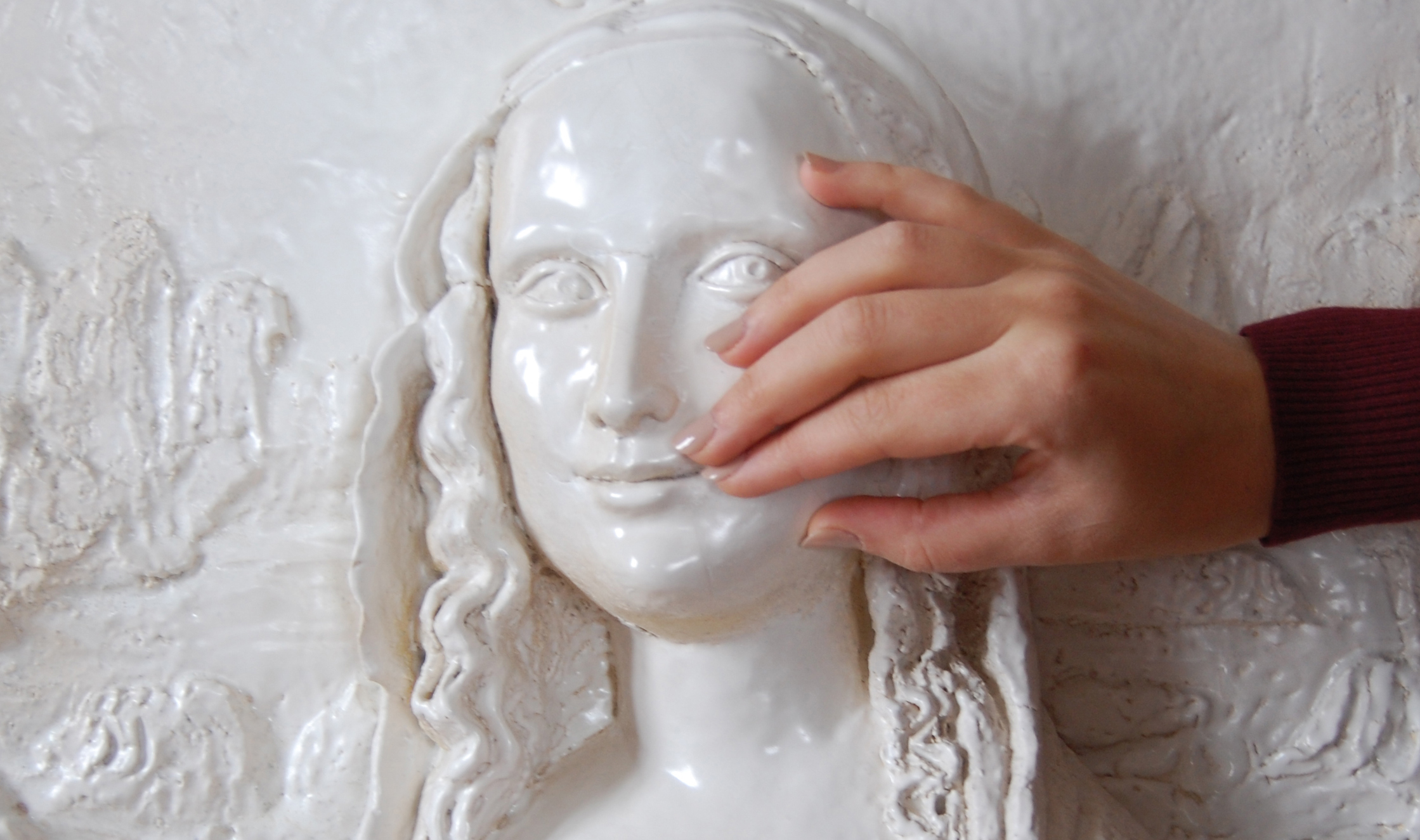 Bas-relief blanc brillant du visage et du cou d’une jeune femme aux traits réguliers, le regard tourné vers la droite, avec un léger sourire et de long cheveux ondulés. Une main touche la joue du bas-relief.