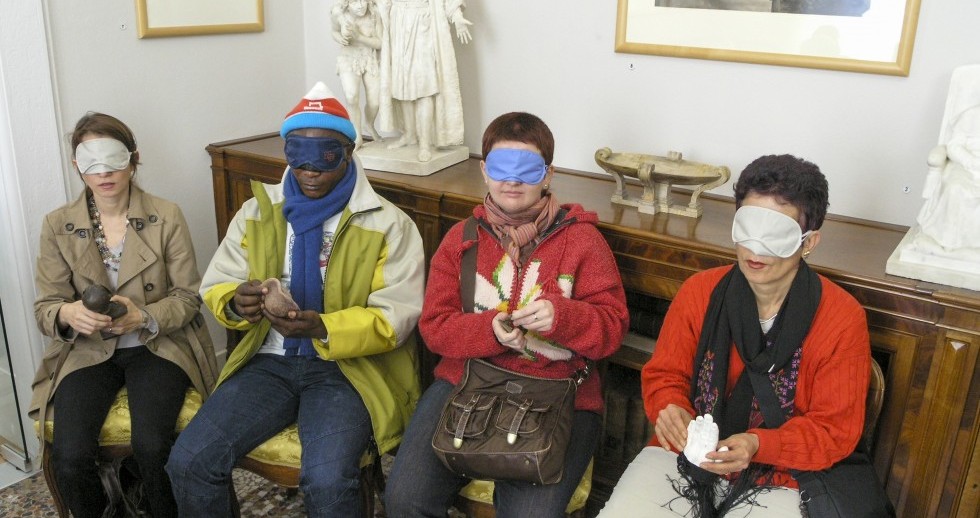 Quattro persone con gli occhi bendati tastano un oggetto che tengono tra le mani. Sono sedute l’una accanto all’altra su antiche sedie in tessuto broccato. Sul mobile intarsiato alle loro spalle ci sono statuette di marmo. 