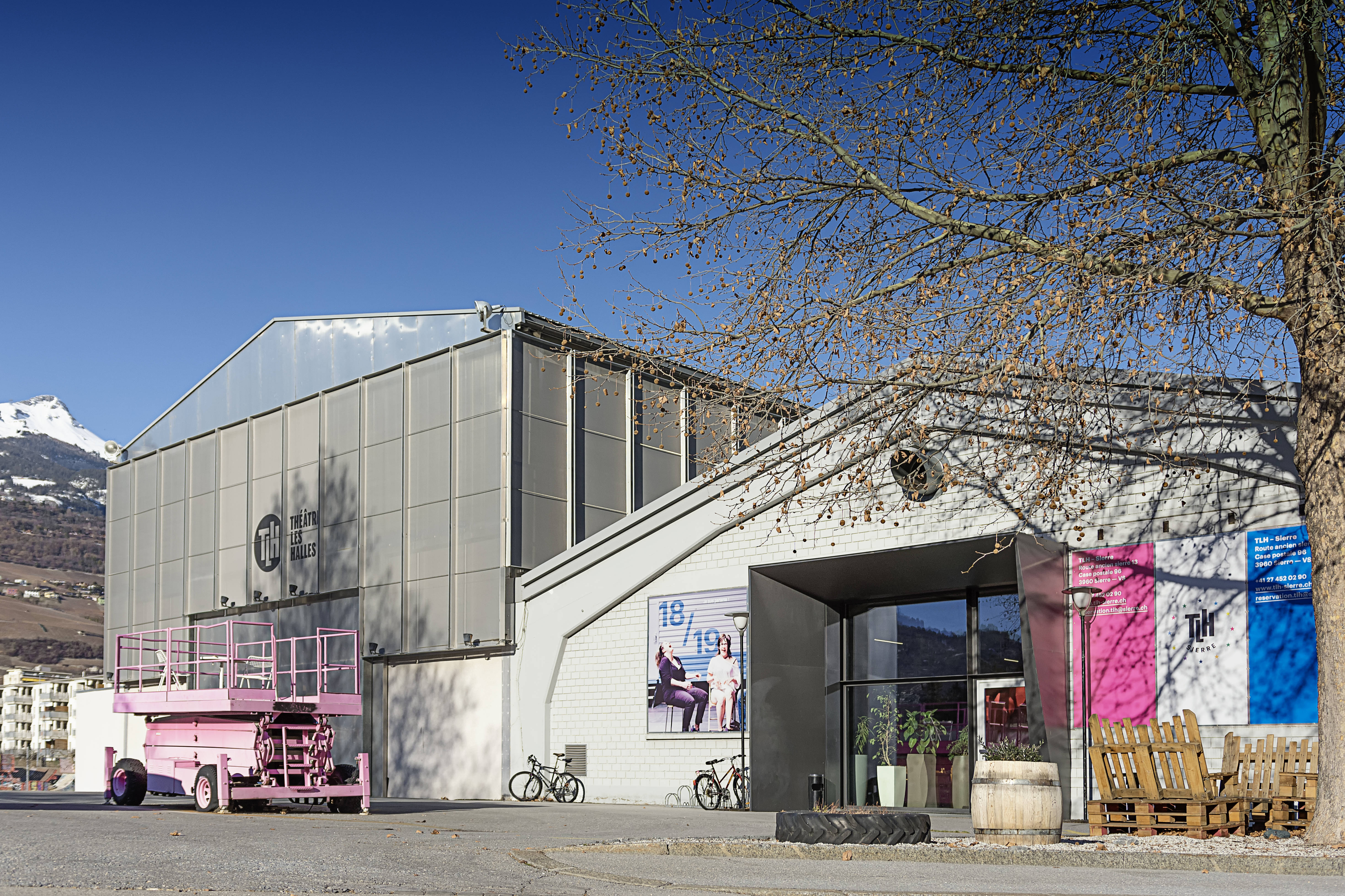 Deux bâtiments sans charme dans une zone industrielle : un entrepôt avec le logo THL et devant un élévateur rose sur roues. À côté, un bâtiment avec un auvent métallique rectangulaire, une entrée vitrée et de grandes affiches colorées sur les côtés. 