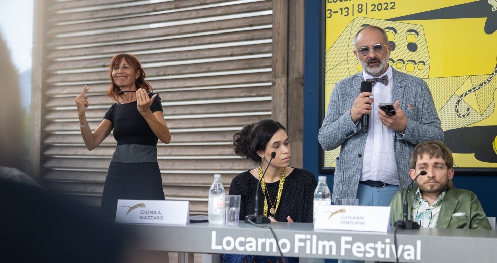 Zwei Personen sitzen an einem Tisch auf dem «Locarno Film Festival» prangt, Mikrofone und Namensschildern vor ihnen. Zwischen ihnen steht ein Mann mit Mikrofon in der Hand. Dahinter hängt das Festivalplakat. Links gebärdet eine schwarzgekleidete Frau. 
