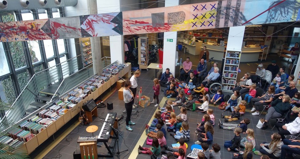 Eine Gruppe in einem Raum mit natürlichem Licht. Zwei Erwachsene stehen neben einem elektronischen Klavier und vor Büchertrögen. Rechts sitzen Kinder und Jugendliche am Boden und schauen die Erwachsenen an. Eine gelbe Linie trennt beide Gruppen. 