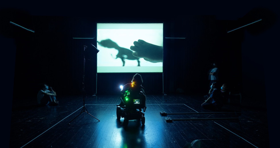 Une personne en fauteuil roulant électrique sur une scène. Dans le fond, une projection vidéo montrant une main et une forme indéfinie. Aux bords de la scène, dans l’obscurité, des personnes regardent la projection.