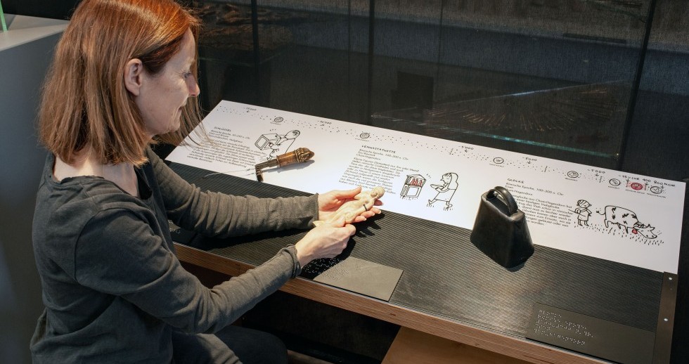 Eine Frau sitzt an einem Tisch, die Kopie einer römischen Statuette in den Händen. Zwei weitere Objekte sind auf der Ablage. Auf der oberen Tischhälfte sind dazu je ein kurzer Text und eine einfache Illustration, unterhalb Braille-Texte angebracht.
