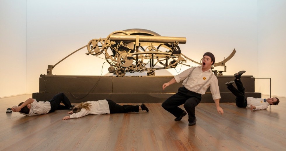 Vier Personen befinden sich vor einem etwa vier Meter breiten Kunstobjekt aus Metall, das auf einem dunkeln Sockel steht. Drei Personen liegen auf dem Boden, eine ist am Fallen.