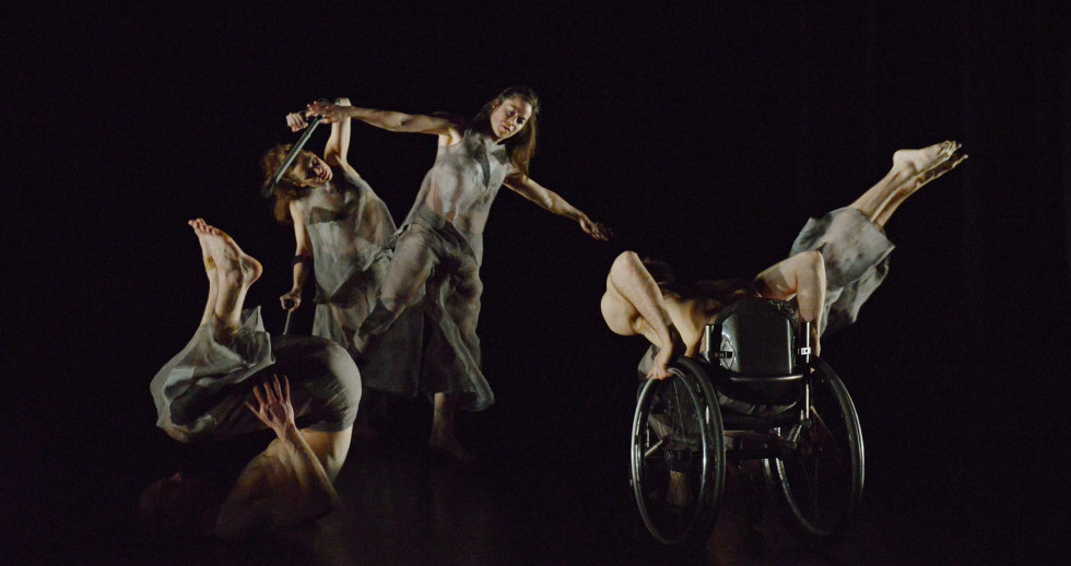 Fünf Menschen barfuss auf einer dunklen Bühne: Balancierend auf einem Bein, sich abstützend auf einer Krücke, mit Kopf, Schultern und Oberarmen auf dem Boden, vorübergebeugt auf einem Rollstuhl sitzend oder die Beine in die Luft streckend.