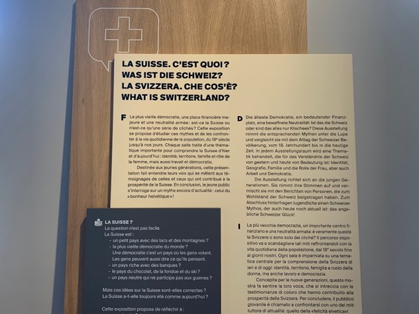 Un panneau avec trois blocs de textes noirs sur fond blanc et un texte blanc sur fond noir. Le titre « La Suisse. C’est quoi ? » est écrit en français, allemand, italien et anglais. En haut du panneau, le contour d’un écusson avec une croix. 