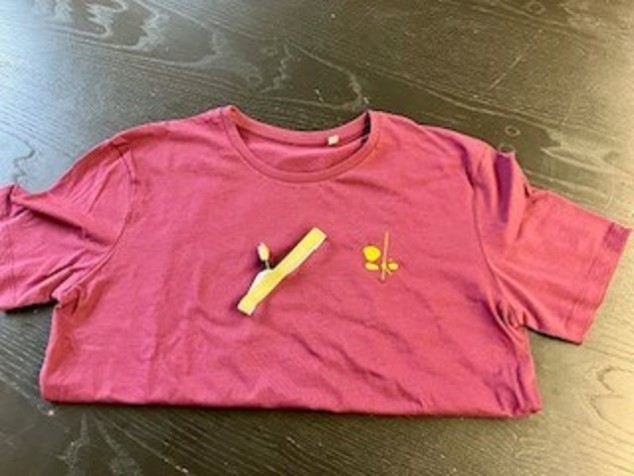 Ein T-Shirt in bordeauxrot mit einer gelben Stickerei. Die Stickerei zeigt abstrakt drei Steine und einen Holz-stab. Auf dem T-Shit liegt ein goldenes Armband mit dem Schriftzug Förderbar. 