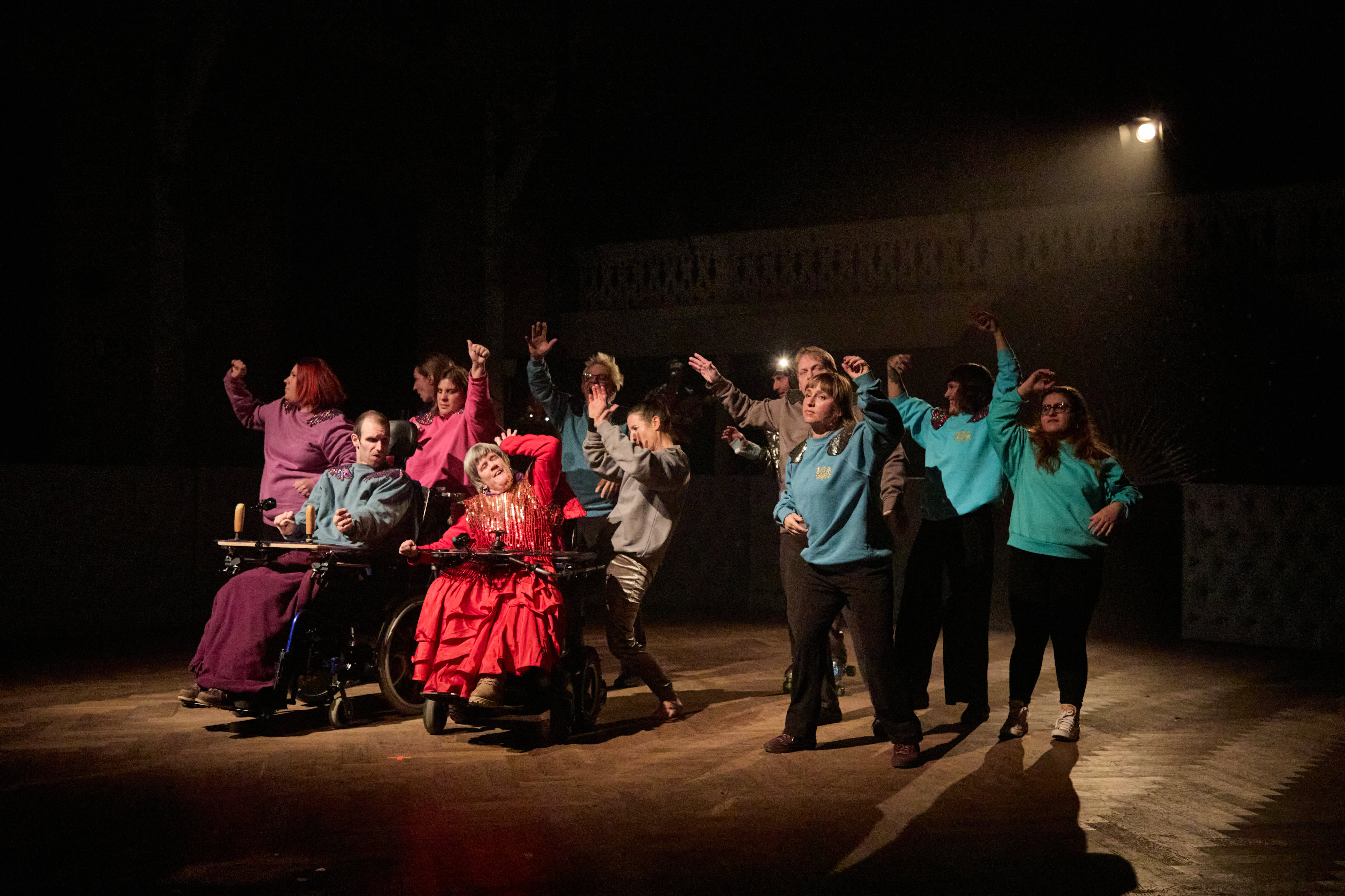 [Translate to Italienisch:] 12 Personen stehen auf einen Holzboden unter Scheinwerferlicht. Zwei Personen sind im Rollstuhl. Der Rest steht. Alle tanzen mit ausgestreckten Armen. 