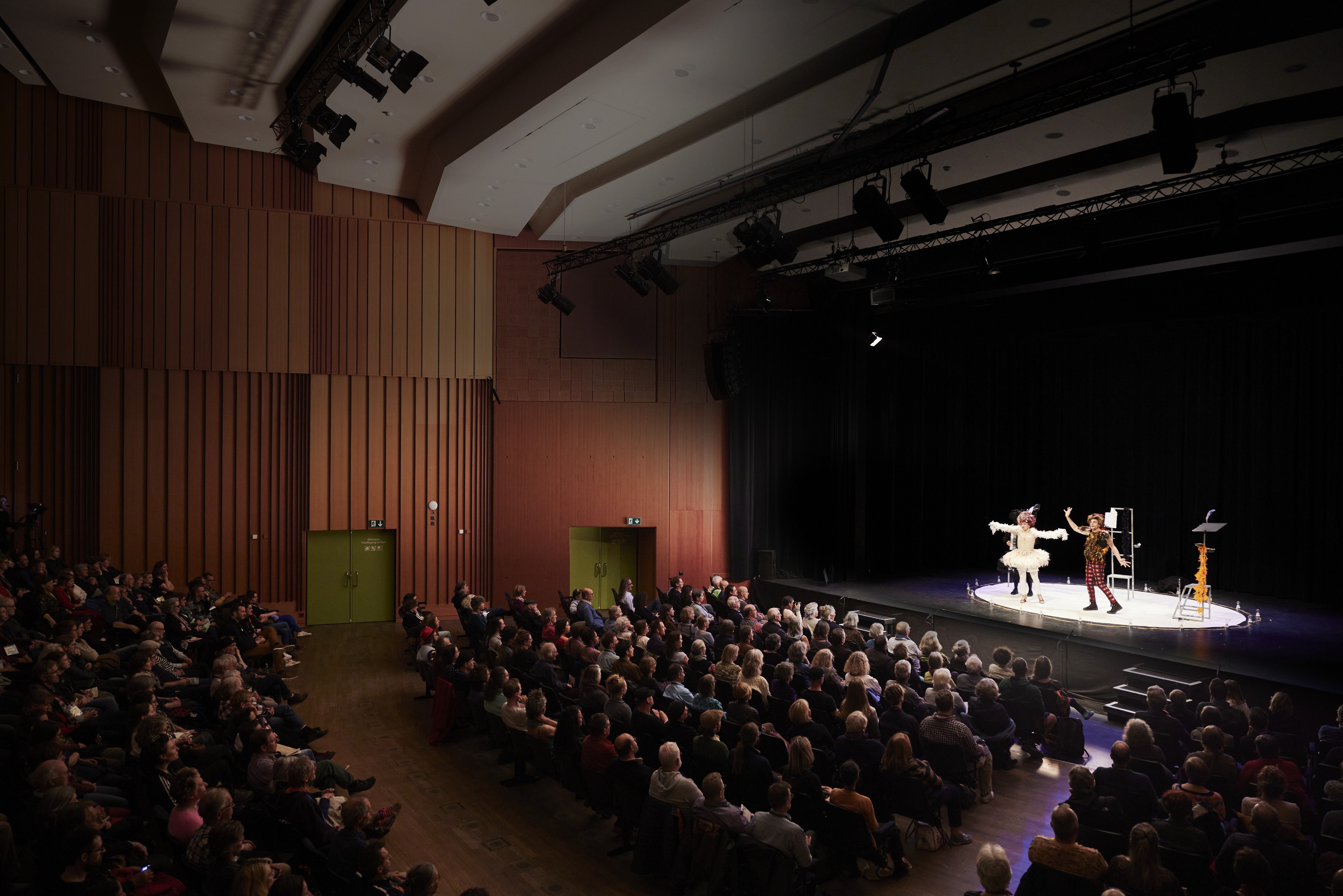 In einem Saal mit vielen hockenden Zuschauern stehen zwei verkleidete Performer auf einer runden weissen Bühne. Die eine Person ist ganz in weiss angezogen, die andere in Karomuster. Sie gestikulieren.