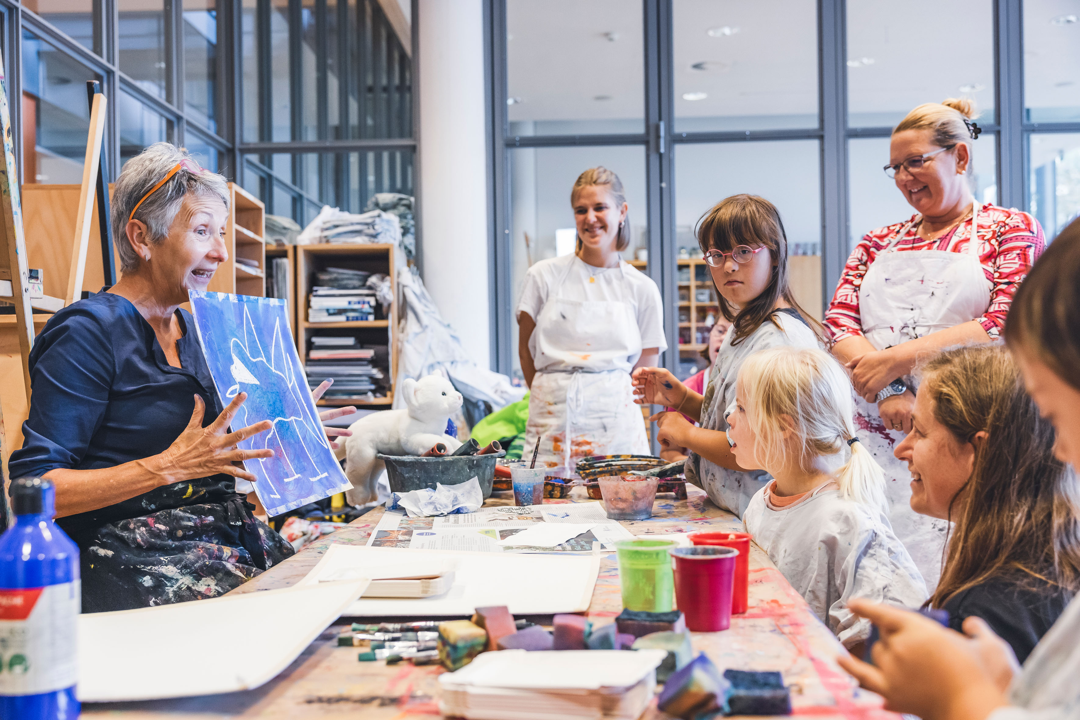 Eine Frau zeigt ein Bild und erklärt etwas. Das Bild ist blau. Kinder und Erwachsene stehen um einen Tisch. Viele lächeln. Ein Kind schaut in die Kamera. Eine Frau zeigt ein Bild und erklärt etwas. Das Bild ist blau. Kinder und Erwachsene stehen um einen Tisch. Viele lächeln. Ein Kind schaut in die Kamera.
