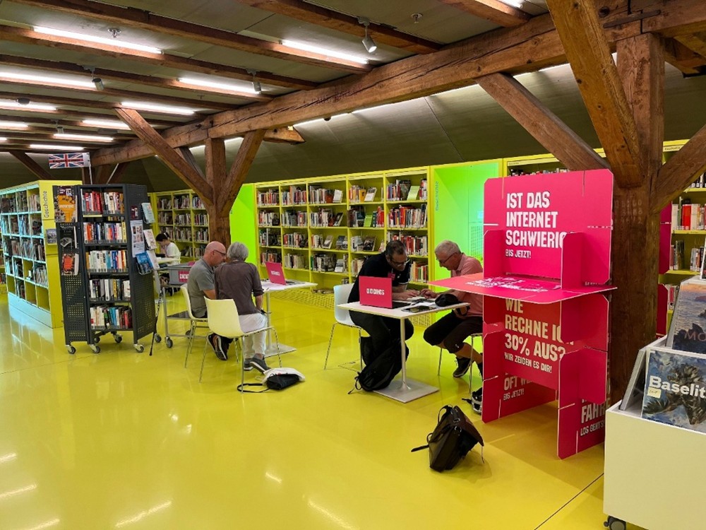 Je zwei Personen sitzen an zwei Tischen. An einem Tisch zeigt eine Person einer anderen etwas auf dem Smartphone. Auf dem Tisch ist zudem ein Laptop aufgeklappt. Rechts steht ein Aufsteller in pinker Farbe mit den Fragen: «Ist das Internet schwierig?» und «Wie rechne ich 30% aus?».