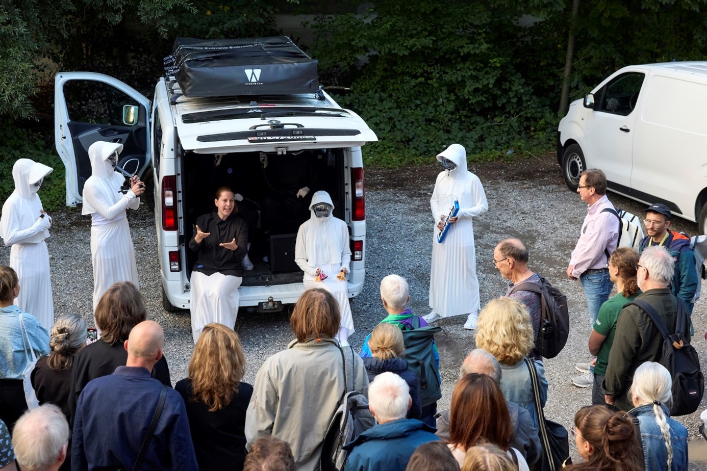 Eine gebärdende Person sitzt auf der Ladefläche eines weissen Vans am Waldrand. Neben ihr stehen in langen weissen Gewändern mit Kapuze gekleidete Personen mit Instrumenten, die Gesichter verborgen. Eine Gruppe von Menschen schaut zu ihnen.