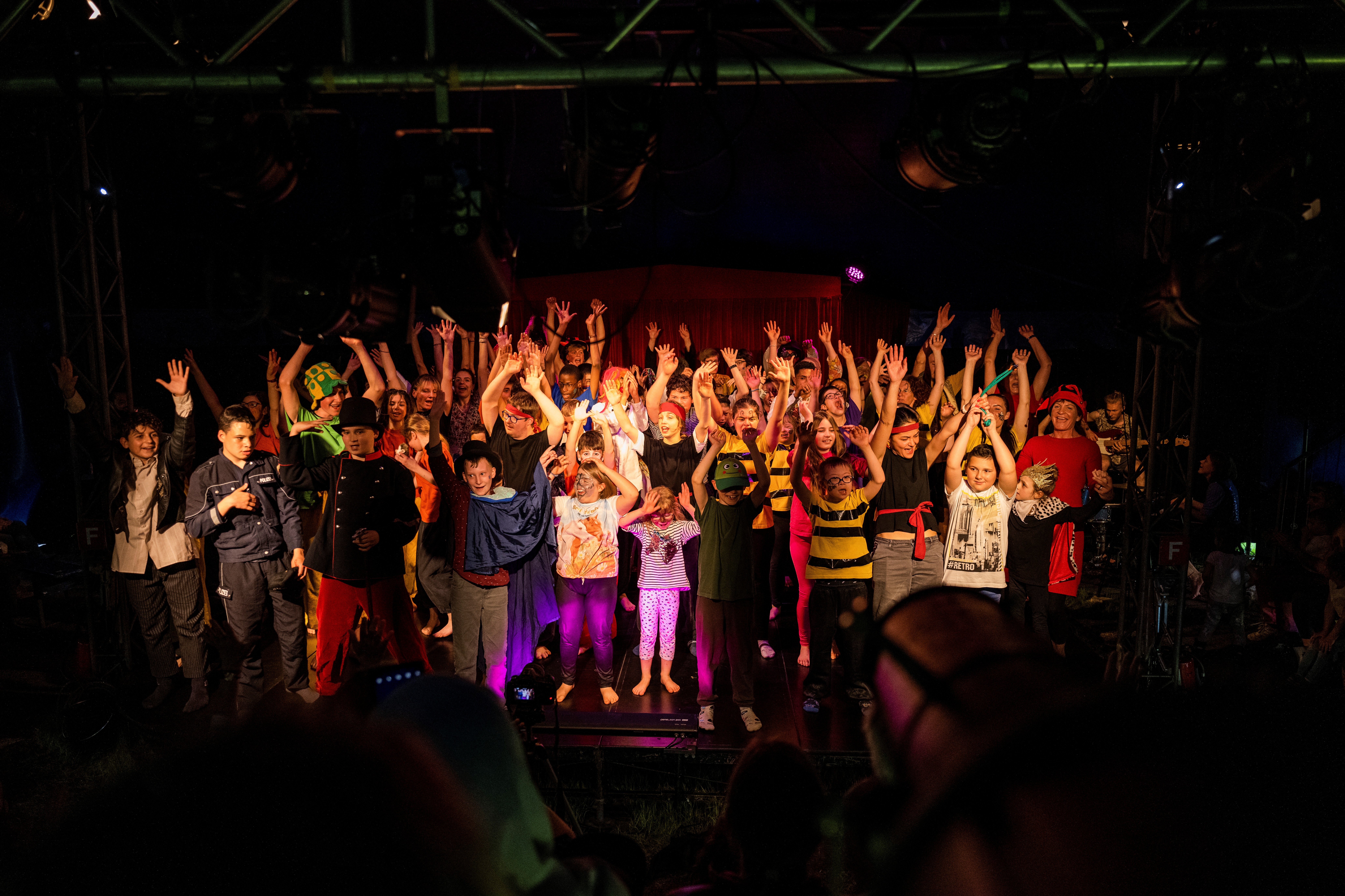 [Translate to Französisch:] Auf einer Bühne stehen etwa 20 Kinder, gross und klein, bunt bekleidet, und halten lachend die Hände in die Luft. Im Vordergrund sind ein paar Hinterköpfe aus dem Publikum zu sehen.