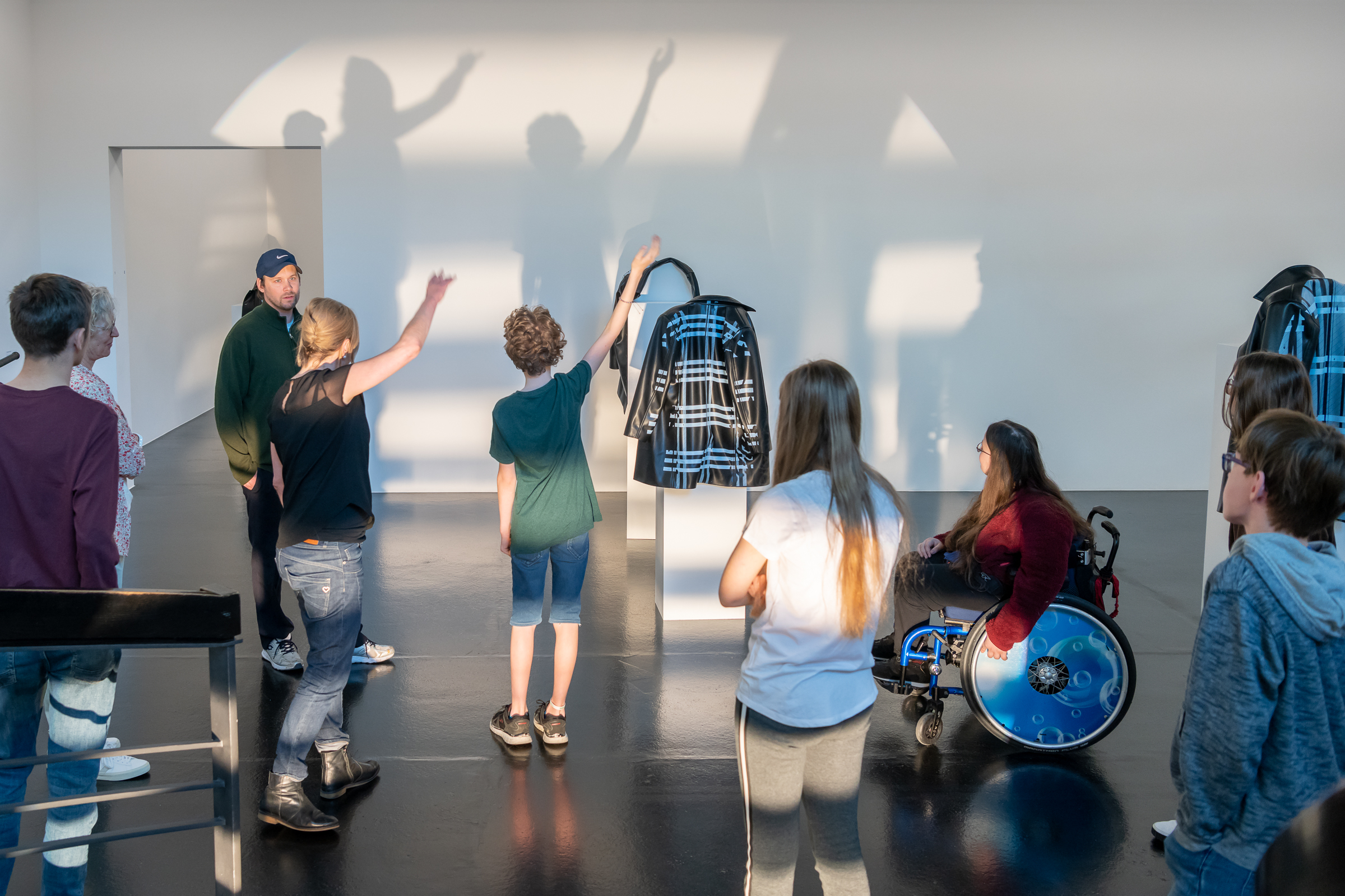 Workshop Situation im Aargauer Kunsthaus. Jugendliche mit und ohne Rollstuhl betrachten ein Kunstwerk in Form einer Jacke und machen Schattenfiguren an die Wand