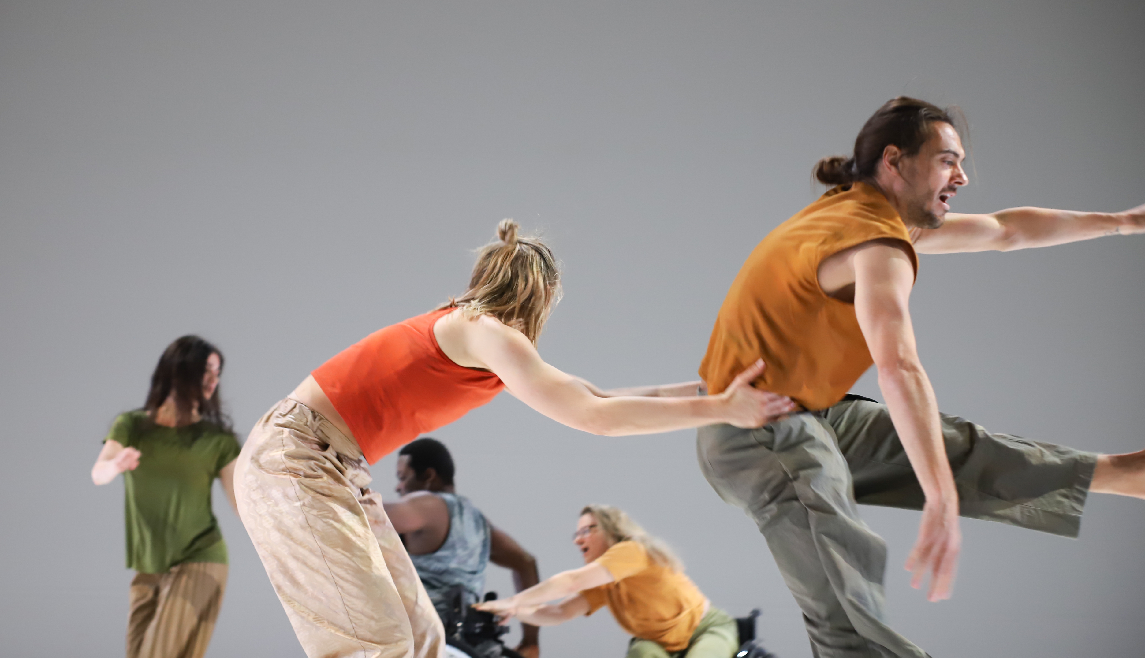 [Translate to Italienisch:] Vor grauem Hintergrund sind fünf Personen aktiv in Bewegung. Im Hintergrund fässt eine blonde Frau im Rollstuhl an den Rollstuhl eines Mannes mit kurzen Haaren vor ihr. Eine weitere Frau in einem grünen T-Shirt dreht sich zu ihnen um. Im Vordergrund packt eine Frau in rotem T-Shirt einem Mann vor ihr in orangem T-Shirt an seiner Hüfte, während er in einem Sprung in der Luft ist.