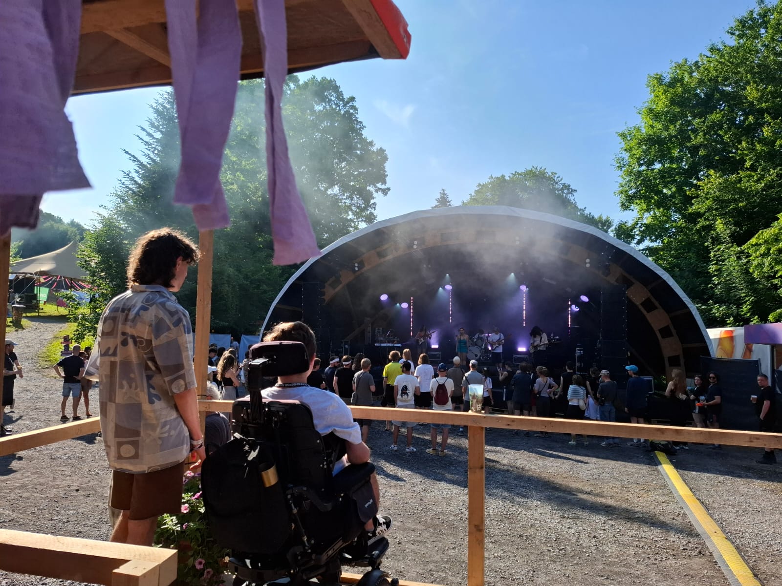 [Translate to Französisch:] Im Vordergrund links ist eine Person stehend und eine Person im Rollstuhl zu sehen. Sie stehen auf einer erhöhten Tribüne mit freier Sicht auf die Hauptbühne. Im Hintergrund steht die Hauptbühne, das Publikum tanzt im strahlend blauen Himmel.