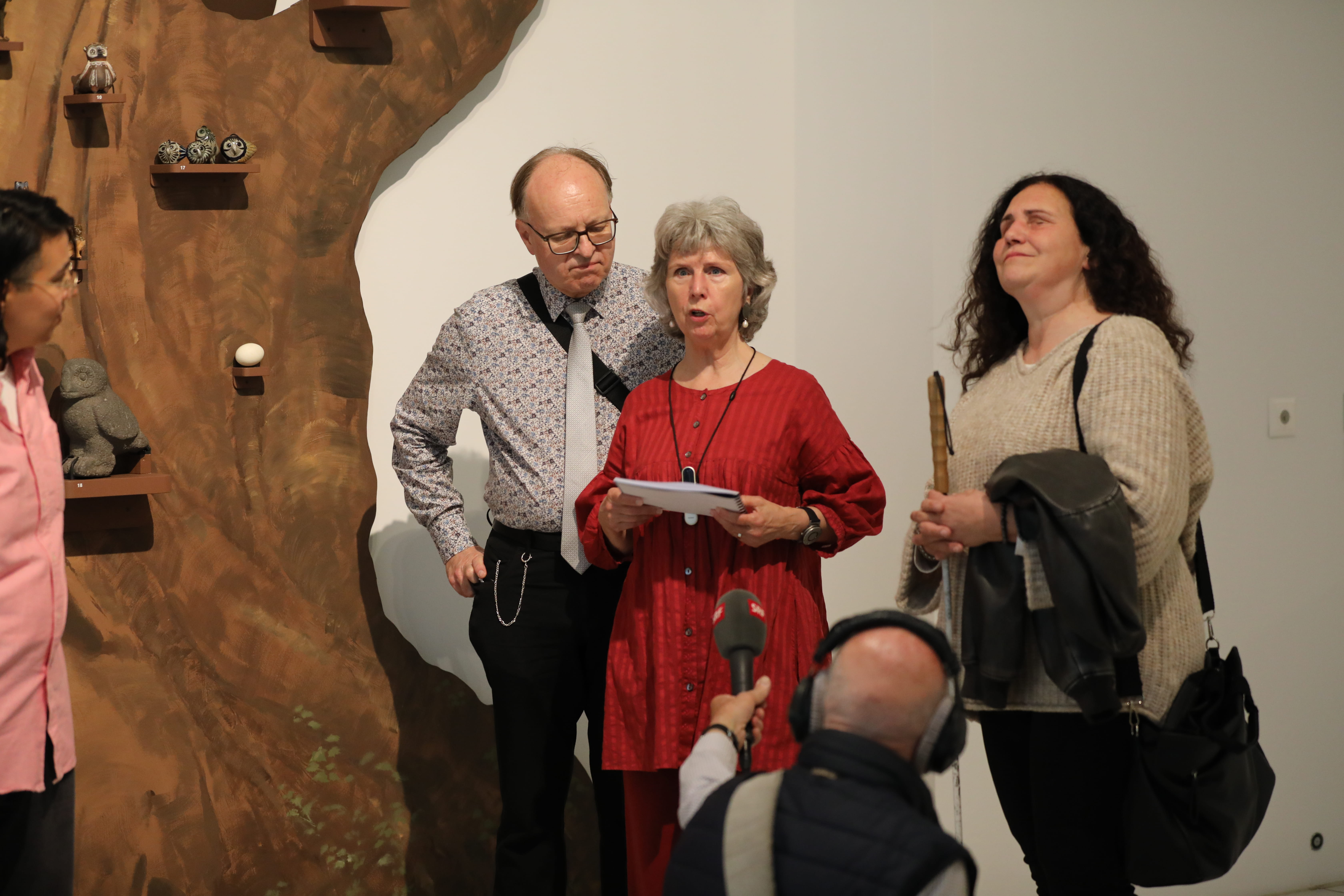 Neben einer Baumskulptur in einem Ausstellungsraum, auf der kleine Eulenfiguren sitzen, stehen ein Mann und eine Frau, die zu zwei weiteren Personen sprechen. Vor ihnen kniet ein Mann mit Mikrofon.