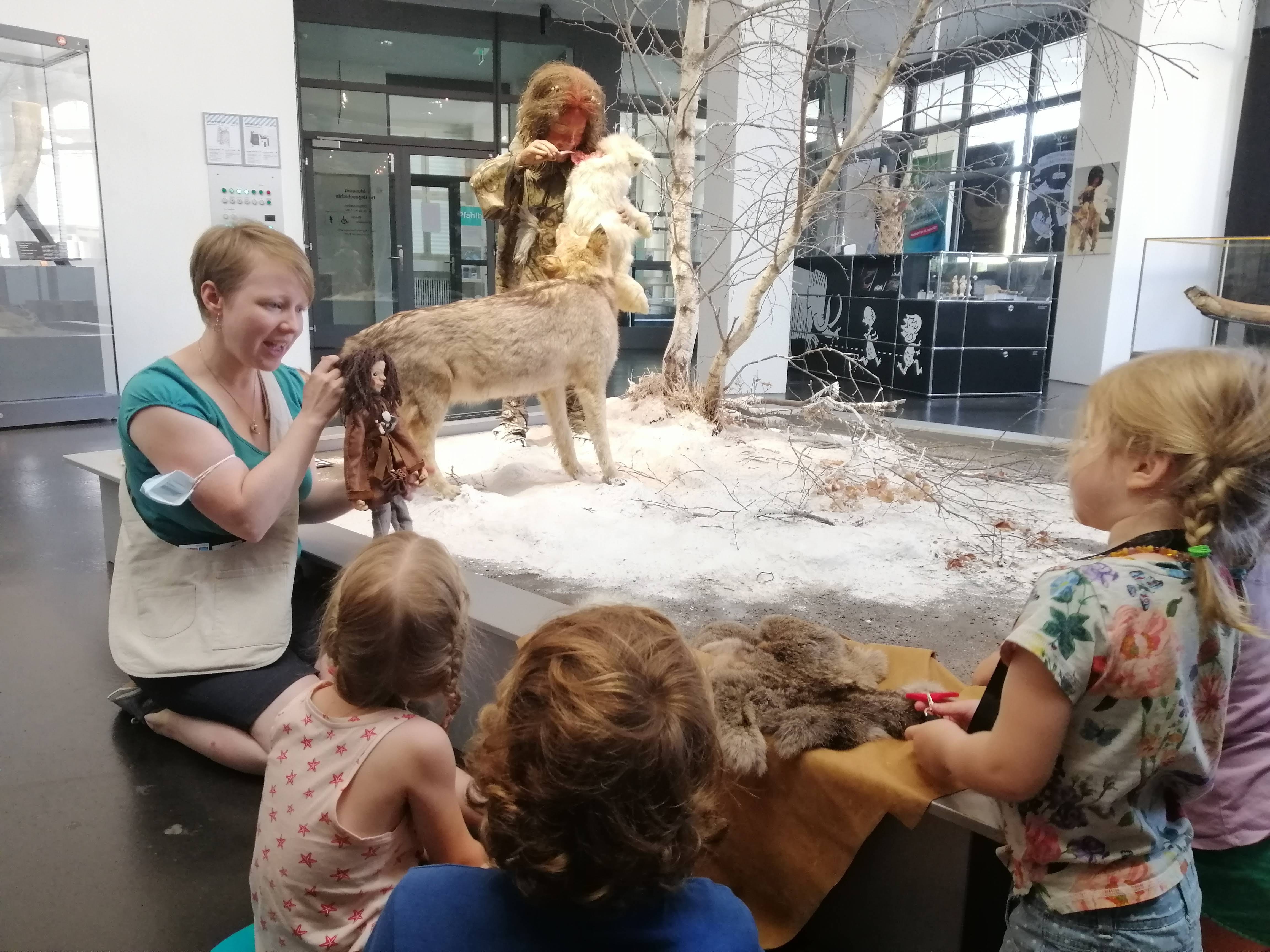 Workshop-Situation im Museum für Urgeschichte(n). Eine Frau kniet am Boden und hält eine Handpuppe. Vor ihr sitzen Kinder. Im Hintergrund sieht man die Figur eines Mädchens in Fellkleidung und einen Hund. Workshop-Situation im Museum für Urgeschichte(n). Eine Frau kniet am Boden und hält eine Handpuppe. Vor ihr sitzen Kinder. Im Hintergrund sieht man die Figur eines Mädchens in Fellkleidung und einen Hund.