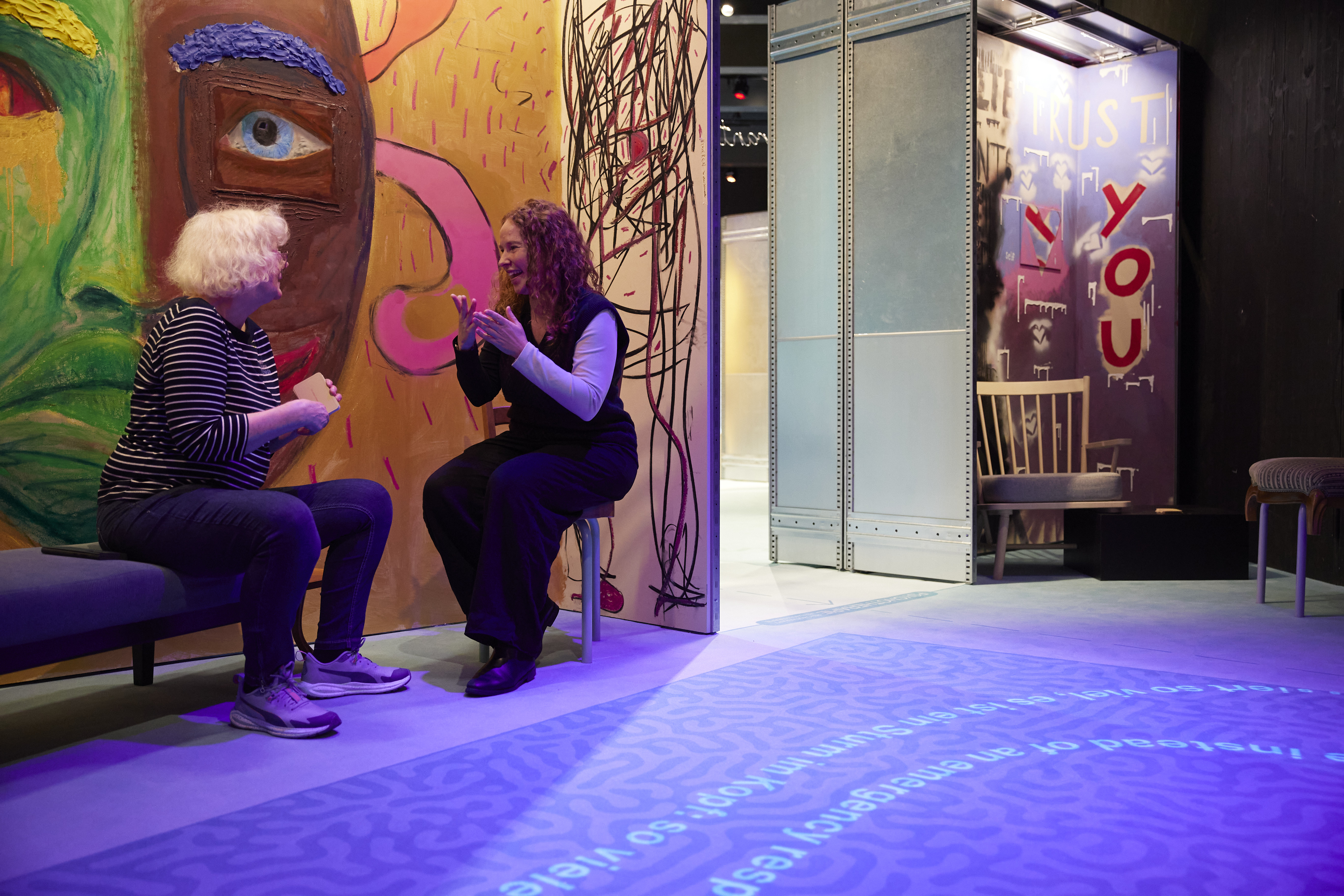 In einem farbigen Ausstellungsraum sitzen sich zwei Menschen gegenüber. Die Person links sitzt auf einem Sofa. Die Person rechts sitzt auf einem Hocker und gestikuliert mit den Händen. Auf der Wand hinter ihnen ein grosses, gemaltes Gesicht.